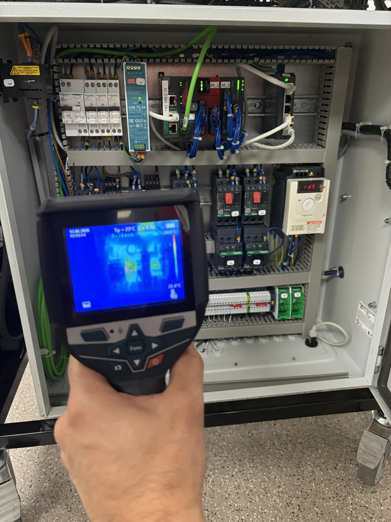 Caméra thermique en main devant un coffret électrique ouvert, image infrarouge visible à l'écran